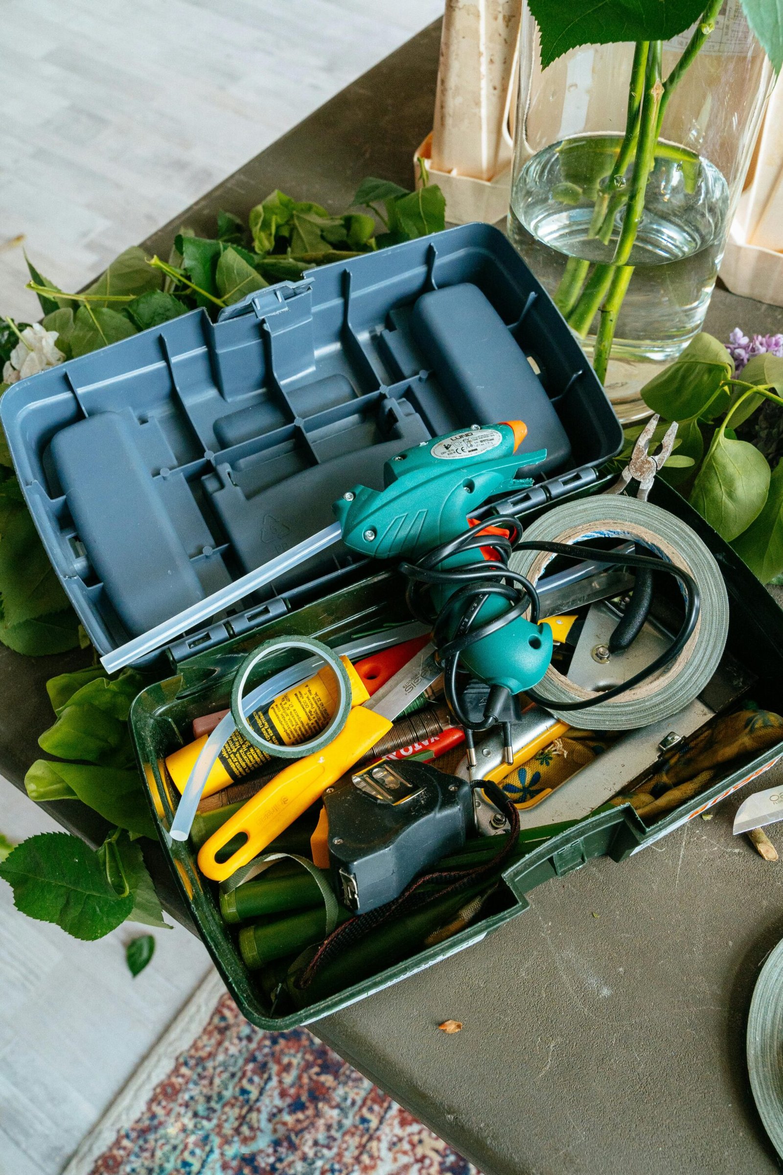 A detailed view of an open toolbox filled with hand tools and a glue gun, surrounded by indoor plants.
